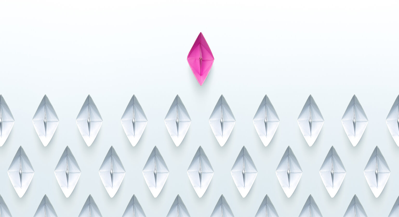 Many paper boats from an overhead point of view. All but one, the boat leading the other boats, are white. The lead boat is red.